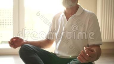 老人在家打坐。 一个成熟的人坐在地板上，坐着一只阿萨纳，双手交叉在膝盖上，阳光明媚
